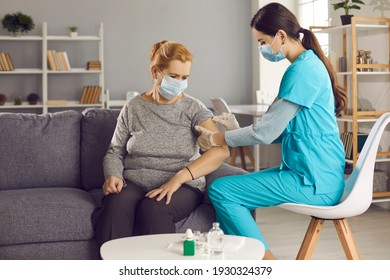 Safe Vaccination For Old People. Mature Woman In Medical Face Mask Getting Flu Or Covid-19 Vaccine Sitting On Sofa At Home. Doctor Or Nurse Giving Flu Or Covid-19 Shot To Senior Female Patient