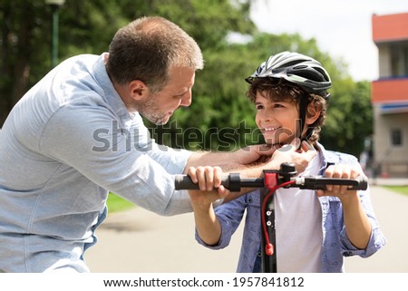 Safety Concept. Close up portrait of smiling adult father putting protection helmet on little boy at park, teaching his brave son to ride electric motorized scooter. Ourdoor Weekend Activity