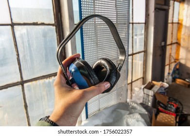 Safety Concept During Work During Production During Machine Noisy. A Man Holds Blue Headphones In His Hands On The Background Of An Old Building Without Repair.