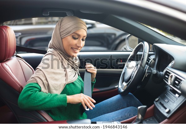 Safety Concept. Young Muslim Woman In Hijab\
Fastening Seat Belt In Car, Smiling Islamic Lady Enjoying Safe\
Drive In Her New Automobile, Making Regulation, Side View Inside\
Vehicle Interior