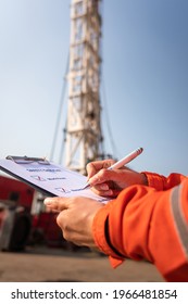 A Safety Office Is Writing On Checklist Document During Perform Safety Audit And Risk Verification At Drilling Site Operation With Blurred Background Of Mount Truck Rig. Selective Focus At Hand.