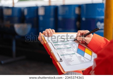 A safety officer is checking on the hazardous material checklist form with chemical storage area at the factory as background. Industrial safety working scene, selective focus.