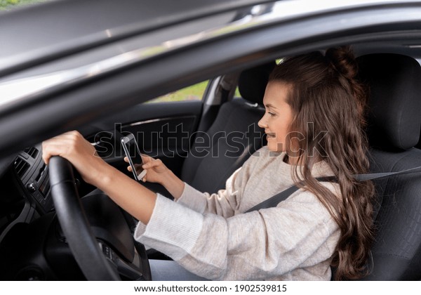 safety and people\
concept - happy smiling young woman or female driver with\
smartphone driving car in\
city