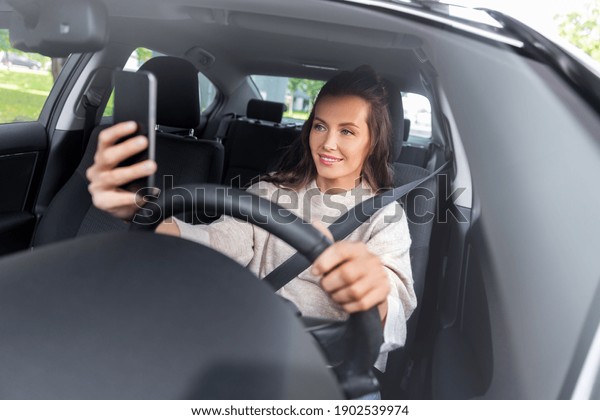 safety and\
people concept - happy smiling young woman or female driver driving\
car and taking selfie with\
smartphone