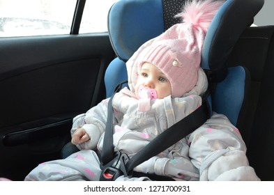 Safety Seat For Baby In Car. Little Cute Girl In A Pink Hat And Overalls Sits Winter In A Child Fastened With Straps.