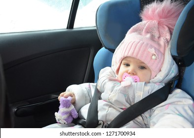 Safety Seat For Baby In Car. Little Cute Girl In A Pink Hat And Overalls Sits Winter In A Child Fastened With Straps.