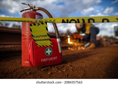 Safety workplace yellow striped caution tape warning sign barricade exclusion zone preventing from public access first aid kit with fire extinguisher defocused construction worker using oxy hot work   - Shutterstock ID 2084357563