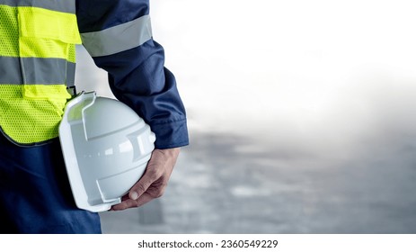 Safety workwear concept. Male hand holding white safety helmet or hard hat. Construction worker man in protective suit and reflective green vest standing with building concrete floor in the background - Powered by Shutterstock