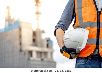 Safety workwear concept. Male hand holding white safety helmet or hard hat. Construction worker man with reflective orange vest and protective gloves standing at unfinished building with tower crane