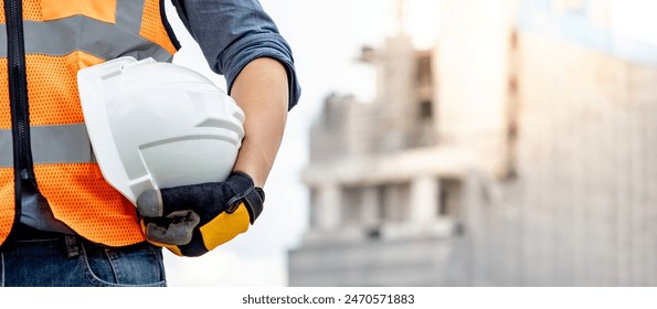 Safety workwear concept. Male hand holding white safety helmet or hard hat. Construction worker man with reflective orange vest and protective gloves standing at unfinished building with tower crane - Powered by Shutterstock