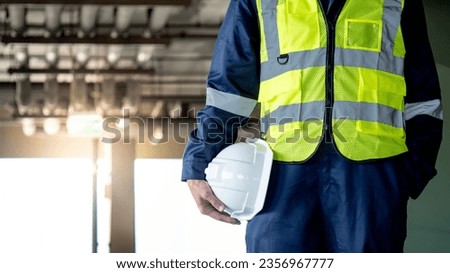 Safety workwear concept. Male hand holding white safety helmet or hard hat. Construction worker man in protective suit and reflective green vest standing with building piping system in the background