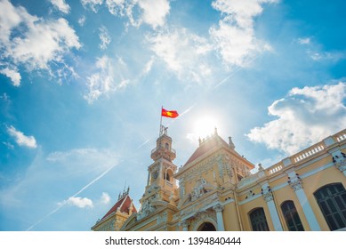 Saigon City Hall With Waving Vietnamese Flag Against Blue Sky And Curly Summer Clouds, Sun, And Airplane Trail. A.k.a. Ho Chi Minh City People's Committee (1900s), A Big Tourist Attraction Of The City