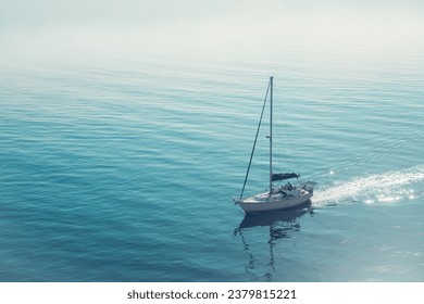 A sail boat gliding across blue water in the early morning sunlight with fog in the background. - Powered by Shutterstock