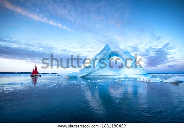 Sail boat with red sails cruising\
among massive ice bergs during dusk. Disko Bay,\
Greenland.
