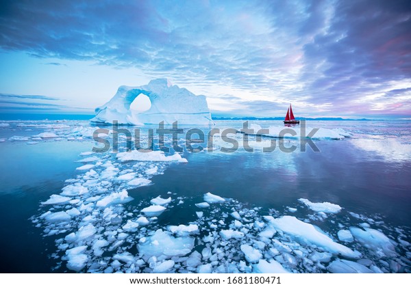 Sail boat with red sails\
cruising next to a pierced ice berg after sunset. Disko Bay,\
Greenland.
