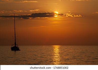 A sailboat anchored at sunset with the sun half hidden by a cloud and the orange coloured horizon - Powered by Shutterstock