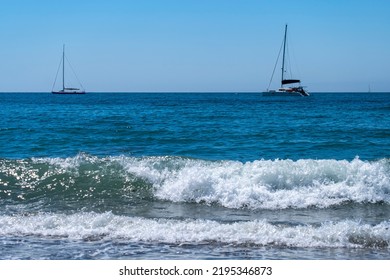 A Sailboat And A Catamaran Anchored Next To Ocean Waves Crashing On Sandy Beach. Sea Waves Breaking On Shore. Nature Splash On Summer Day. Sea Wave Crashing On Beach. Summer Holidays Concept.