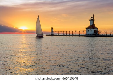 A sailboat rounds the breakwater near sunset on the longest day of the year by the Lighthouses at St. Joseph, Michigan with people fishing from and walking on the pier.