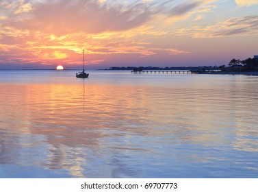 A sailboat silhouetted against a half set sun reflected in the quiet waters of a cove off Pensacola Bay, Florida