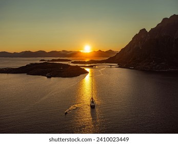 
A sailboat under the midnight sun, behind a large hill in Lofoten, near Henningsvaer, in the distance tall mountains, sun setting.