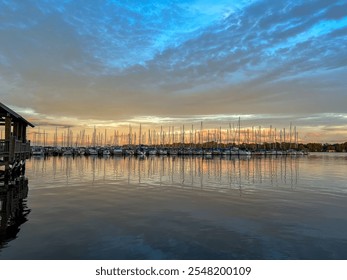 Sailboats docked in a serene marina reflect on the calm water, framed by a warm sunset with vibrant orange and blue hues, under a textured sky filled with soft clouds. - Powered by Shutterstock