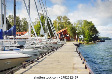 Sailboats Moored To A Pier. Traditional Houses Colored With Falu Red Dye.  Björkö Island, Lake Mälaren, Sweden