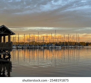 Sailboats rest in a marina at sunset, their masts mirrored in the calm water, under a sky brushed with warm orange and golden hues, framed by a wooden dock on the left. - Powered by Shutterstock