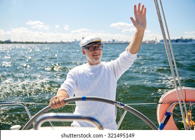 Sailing, Age, Tourism, Travel And People Concept - Happy Senior Man In Captain Hat On Steering Wheel And Waving Hand Sail Boat Or Yacht Floating In Sea