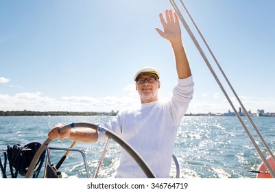 Sailing, Age, Tourism, Travel And People Concept - Happy Senior Man In Captain Hat On Steering Wheel And Waving Hand Sail Boat Or Yacht Floating In Sea