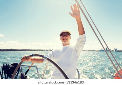 Sailing, Age, Tourism, Travel And People Concept - Happy Senior Man In Captain Hat On Steering Wheel And Waving Hand Sail Boat Or Yacht Floating In Sea