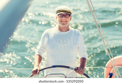 Sailing, Age, Tourism, Travel And People Concept - Happy Senior Man In Captain Hat On Steering Wheel And Navigating Sail Boat Or Yacht Floating In Sea