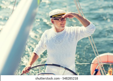 Sailing, Age, Tourism, Travel And People Concept - Happy Senior Man In Captain Hat On Steering Wheel And Navigating Sail Boat Or Yacht Floating In Sea