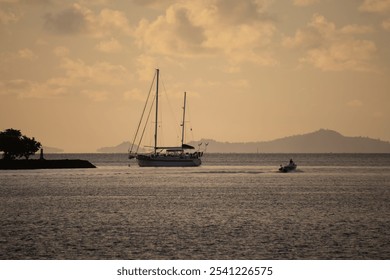 "Sailing boat anchored in calm waters at golden hour, with a motorboat cruising past. Silhouettes of mountains and a tree enhance the serene, golden-lit scene." - Powered by Shutterstock