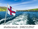 Sailing on the motorboat from the city of Sorvagur to the island of Mykines on Faroe islands. National Faroese flag waving against a cloudy sky. Landscape photography