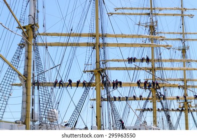 Sailors On The Rigging Of A Tall Ship Balanced Along The Yard Arm Attending To The Sails, Multiple Masts And Crew Against A Blue Sky