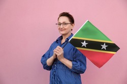 Saint Kitts And Nevis Flag. Woman Holding St Kitts And Nevis Flag. Nice Portrait Of Middle Aged Lady 40 50 Years Old Holding A Large Flag Over Pink Wall Background On The Street Outdoors.