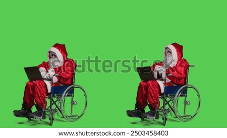 Saint nick with disability using laptop in studio, attending online videocall meeting and browsing webpage. Modern santa character in wheelchair working with internet on portable computer.