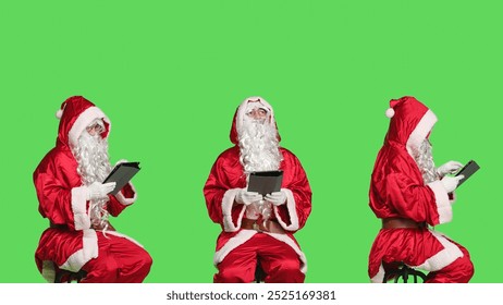 Saint nick using tablet in studio sitting on chair against greenscreen background, working on modern device to create presents list. Modern santa claus browsing website on gadget.