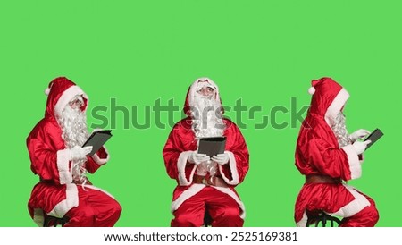 Saint nick using tablet in studio sitting on chair against greenscreen background, working on modern device to create presents list. Modern santa claus browsing website on gadget.