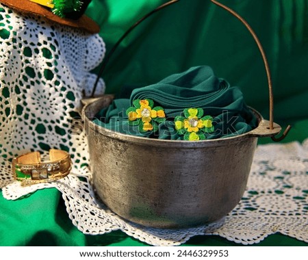 Saint Patrick's Day decoration, still life. Leprechaun's attributes: handmade four-petaled clover leaves (ear-rings) on green canvas in metal cauldron. Irish folklore. White crochet pattern in bg.