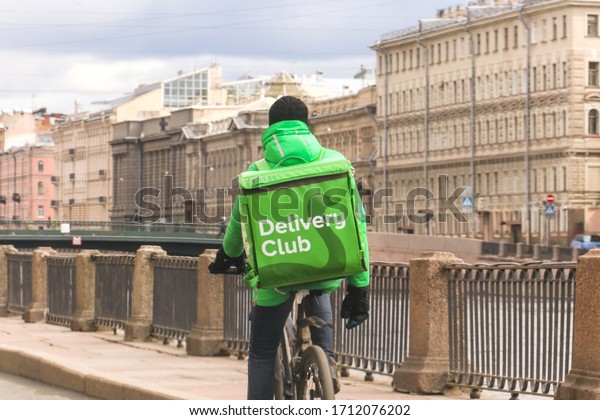 Saint\
Petersburg, Russia, April 18, 2020: a courier in a uniform and\
branded green delivery club backpack rides against parked\
cars.Thermos bag with logo. Food\
delivery.
