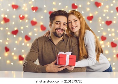 Saint Valentine's Day celebration, gift giving and romantic couple concept. Portrait of happy smiling young man and woman hugging and holding red present box against lights and hearts decor background