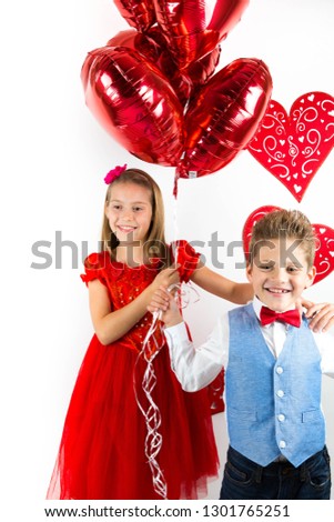 Saint Valentine's day. Pretty girl with red dress and gentleman boy with blue vest, red butterfly tie,  red roses bucket and heart shaped gift box. Valentines day kids. Love and friendship. Woman Day