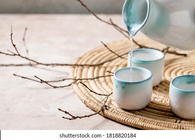 Sake ceramic set for traditional japanese alcohol drink rice wine sake pouring from pitcher in three cups, standing on straw napkin with dry branches over beige texture background.