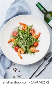 Salad With Arugula, Cherry Tomatoes And Grilled Shrimps In White Porcelain Plate On Light Blue Background With Fork And Knife, Bottle Of White Wine And Blue Napkin. Overhead View, Flat Lay