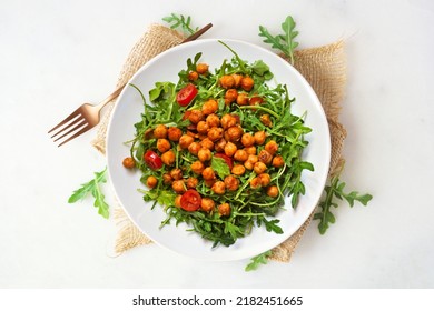 Salad Of Arugula And Seasoned Chickpeas. Overhead View On A Wooden Plate Over A White Marble Background.