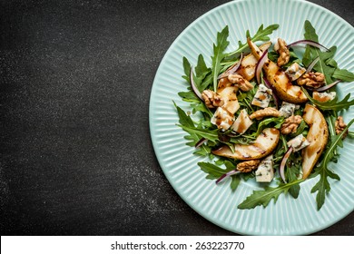 Salad - blue cheese, pear, arugula, walnuts, red onion and balsamic vinegar dressing on pastel blue plate from above. Black chalkboard as background. Layout with free text space.
