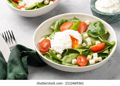 Salad bowl with fresh green spinach, cherry tomatoes, croutons, egg benedict and sour cream dressing on a grey stone surface - Shutterstock ID 2112469361