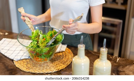 Salad Concept The Woman With White T-shirt And Dark Green Pants Standing At The Kitchen And Making A Bowl Of Green Salad.