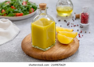 Salad dressing with oil, lemon juice, salt and pepper served in glass cruet or bottle. Close up, ingredients and plate with arugula and tomato salad on background. Horizontal. Basic Vinaigrette.